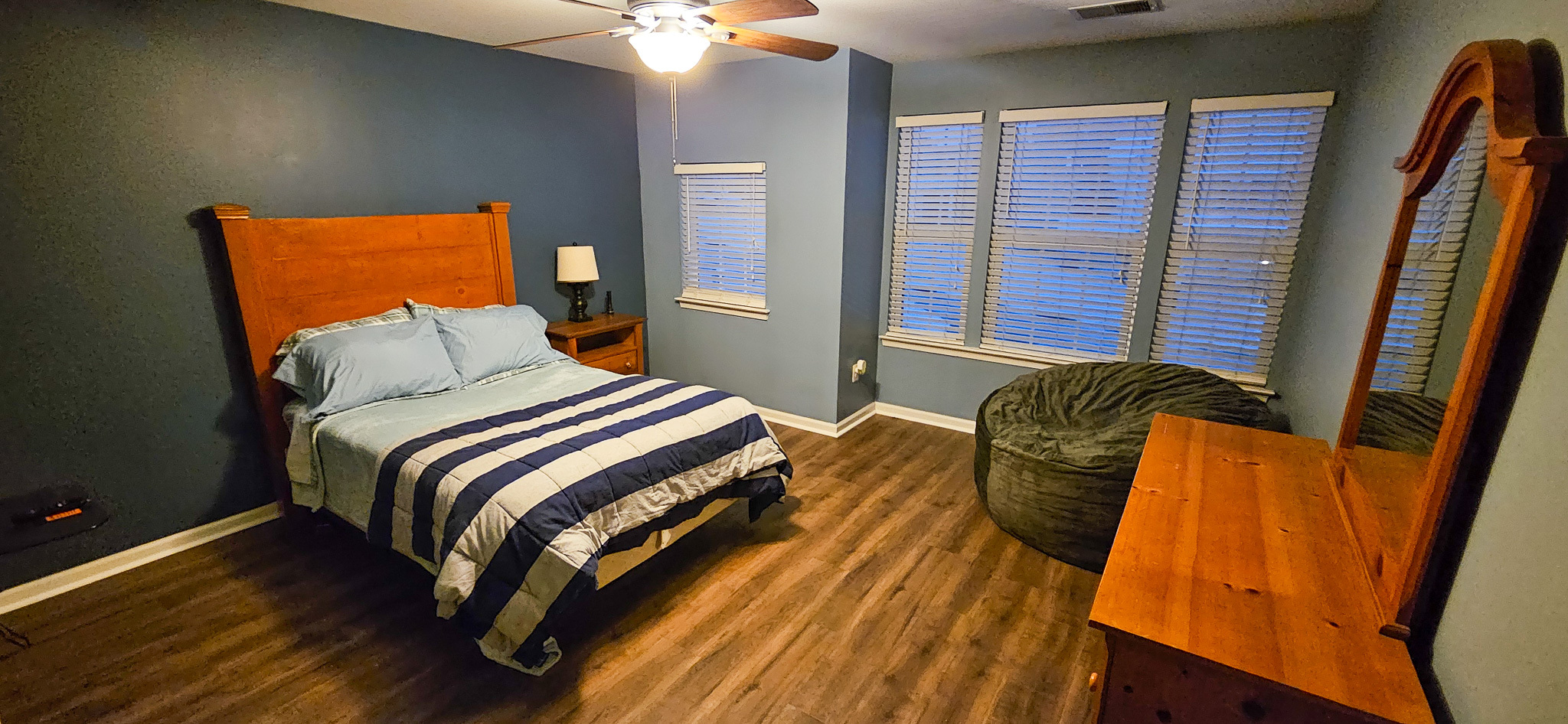A bedroom with a wooden bed frame, blue bedding, a dresser with a mirror, and large windows along one wall inside the home of Sponsored Residential Provider Vanessa Moore in Fredericksburg, Virginia.