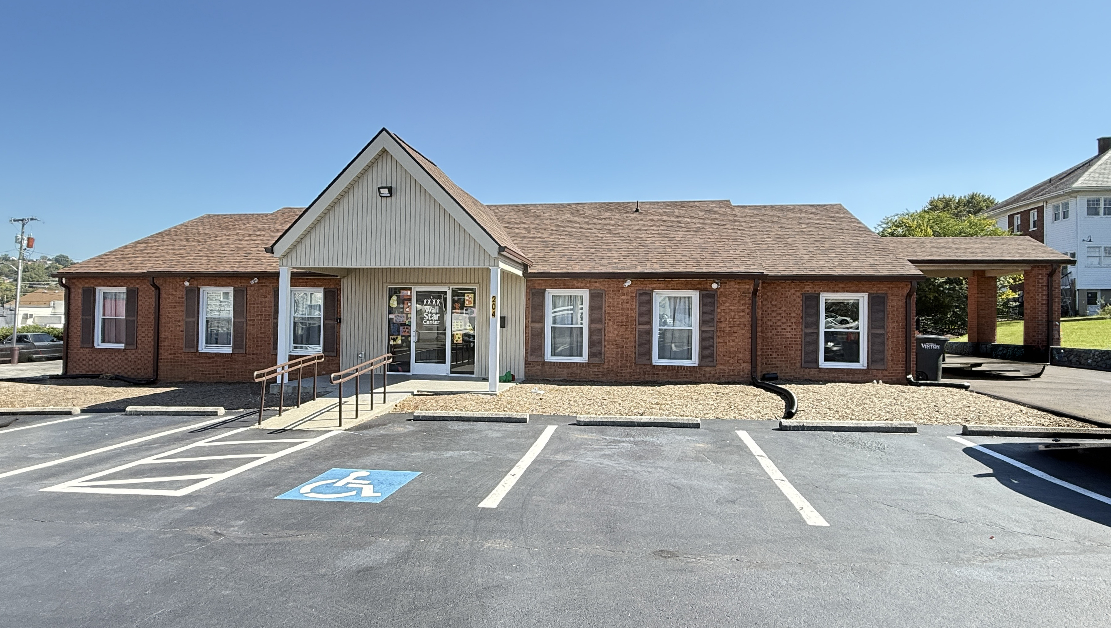 Front entrance of building that houses the Vinton Star Center and Vinton offices for Wall. Brick one story building with covered cream colored porch and main entrance, overhang over drive on the right that connects to back lot, and front parking spaces.