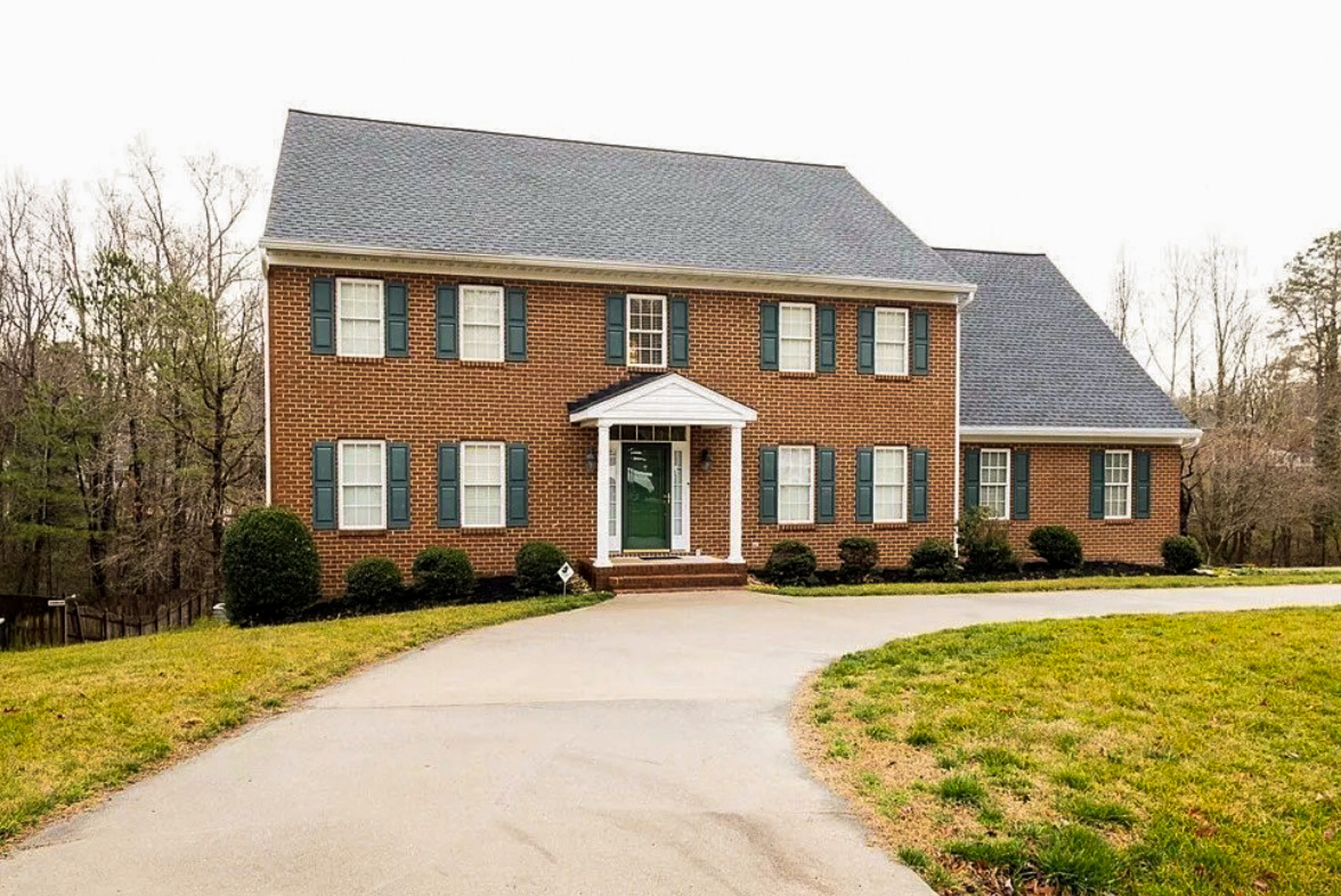 Large two-story brick home with green shutters, a centered front porch with white columns, and a curved driveway in a quiet, wooded setting belonging to Sponsored Residential Provider Wanette and Carol Spencer in Richmond, Virginia.