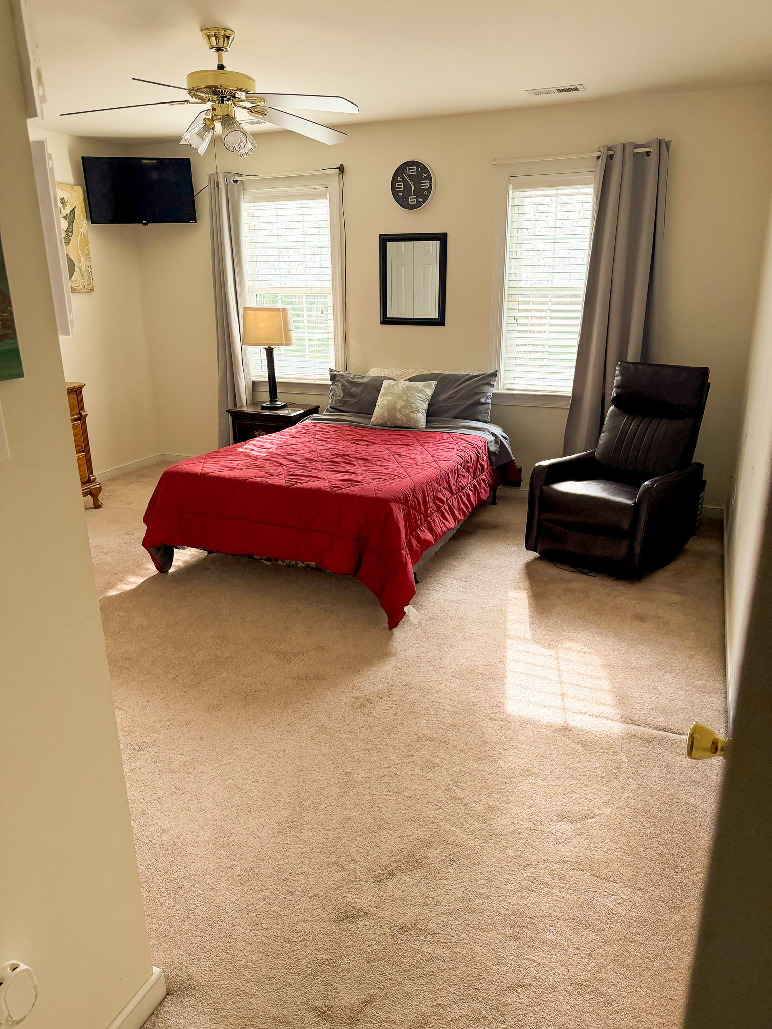 Bright and spacious bedroom with a bed featuring a red comforter, two windows with curtains, a ceiling fan, wall-mounted TV, bedside table with lamp, and a reclining chair belonging to Sponsored Residential Provider Wanette and Carol Spencer in Richmond, Virginia.