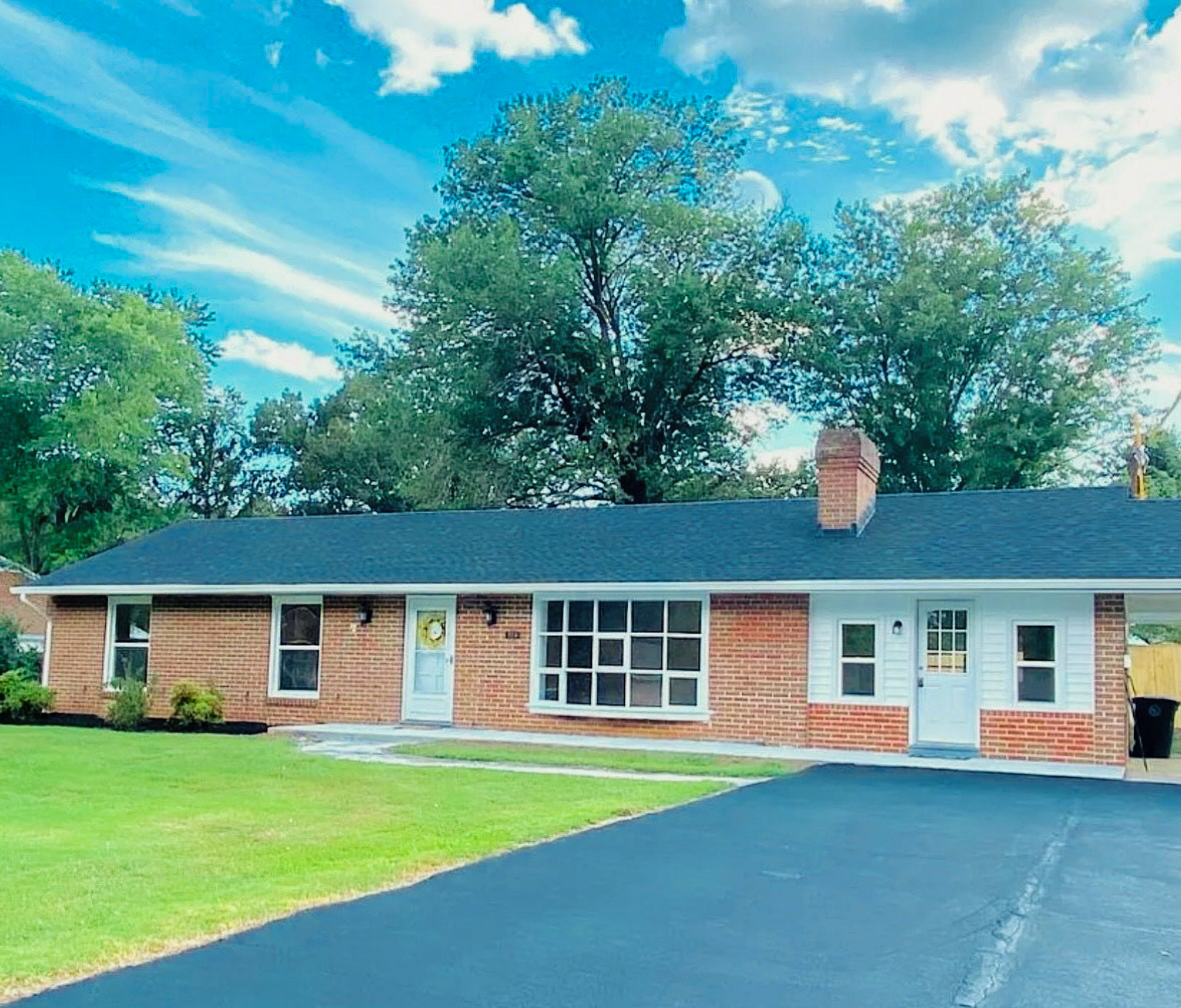 Brick ranch-style home with large front windows, white doors, paved driveway, and green lawn with mature trees belonging to Sponsored Residential Providers Casey and Fred Doswell in Lynchburg, VA.