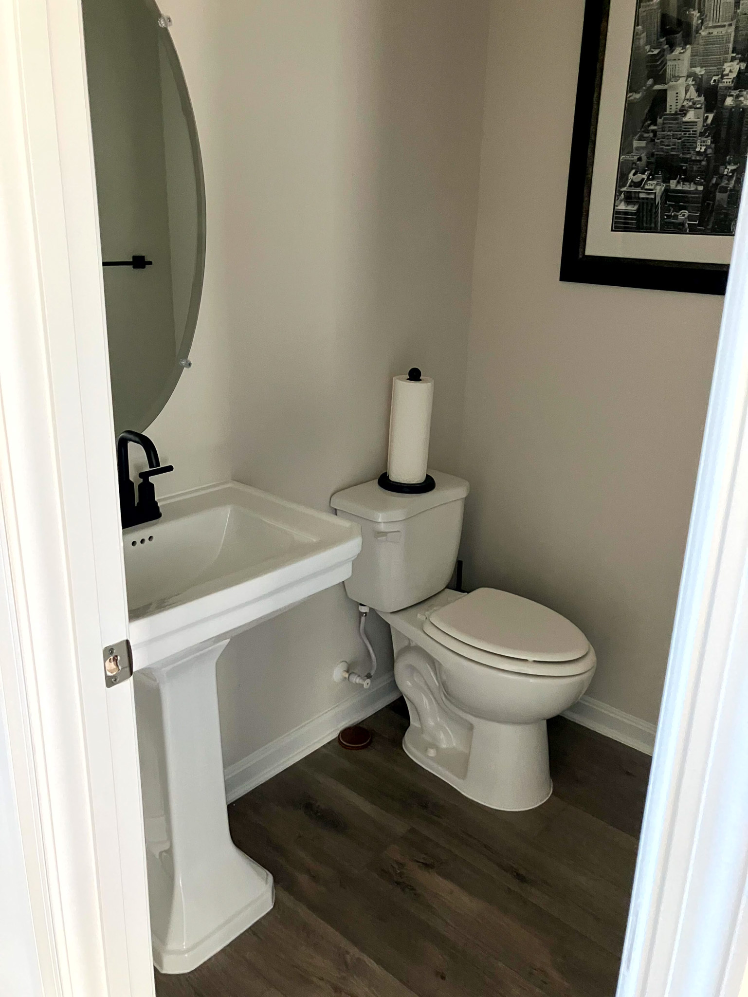 Small bathroom with a pedestal sink, toilet, oval mirror, and framed wall art above wood‑style flooring  inside the home of Sponsored Residential Provider Homaira Yazdani in Fredericksburg, Virginia.