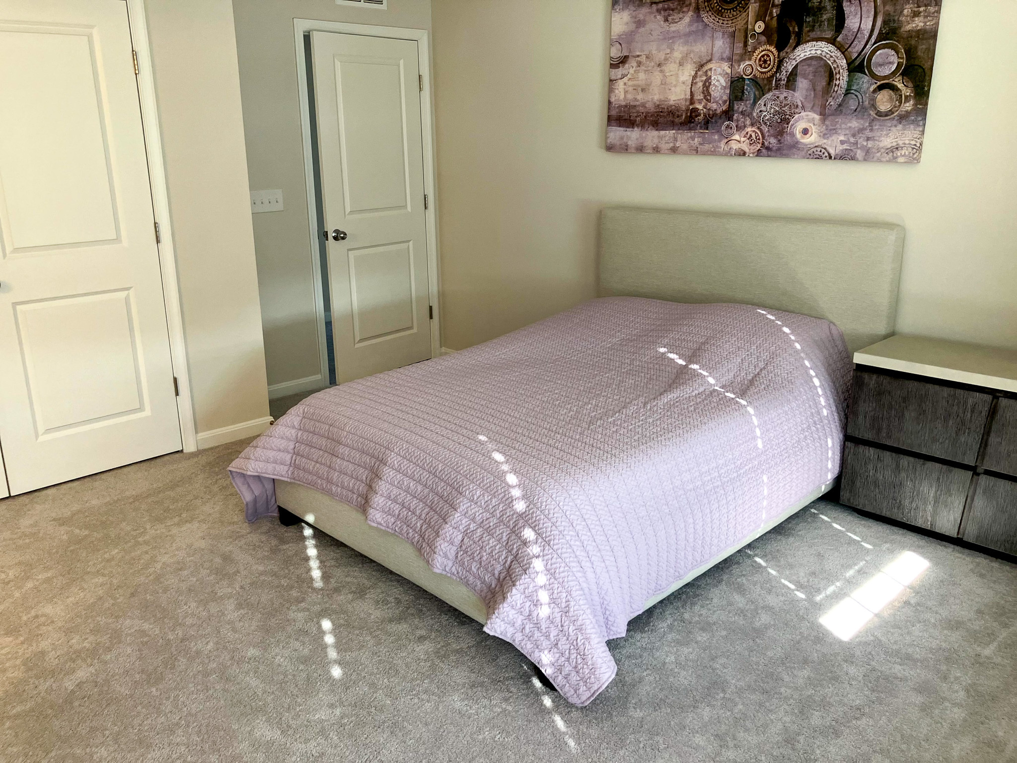 Bedroom with a light‑gray upholstered bed, lavender quilt, bedside dresser, and two closed white doors  inside the home of Sponsored Residential Provider Homaira Yazdani in Fredericksburg, Virginia.
