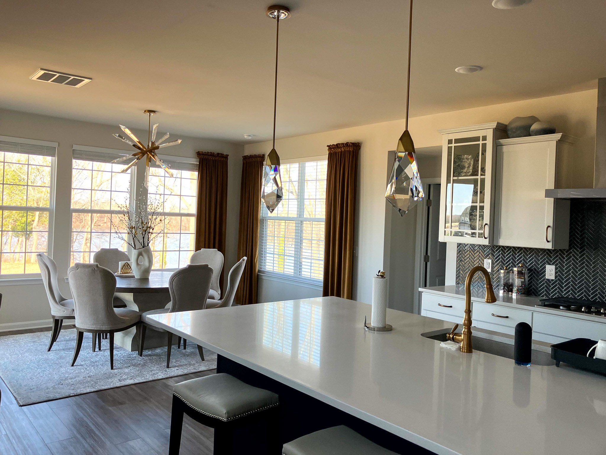 Open kitchen and dining area with a large white island, pendant lights, and a dining table set near tall windows with gold curtains  inside the home of Sponsored Residential Provider Homaira Yazdani in Fredericksburg, Virginia.