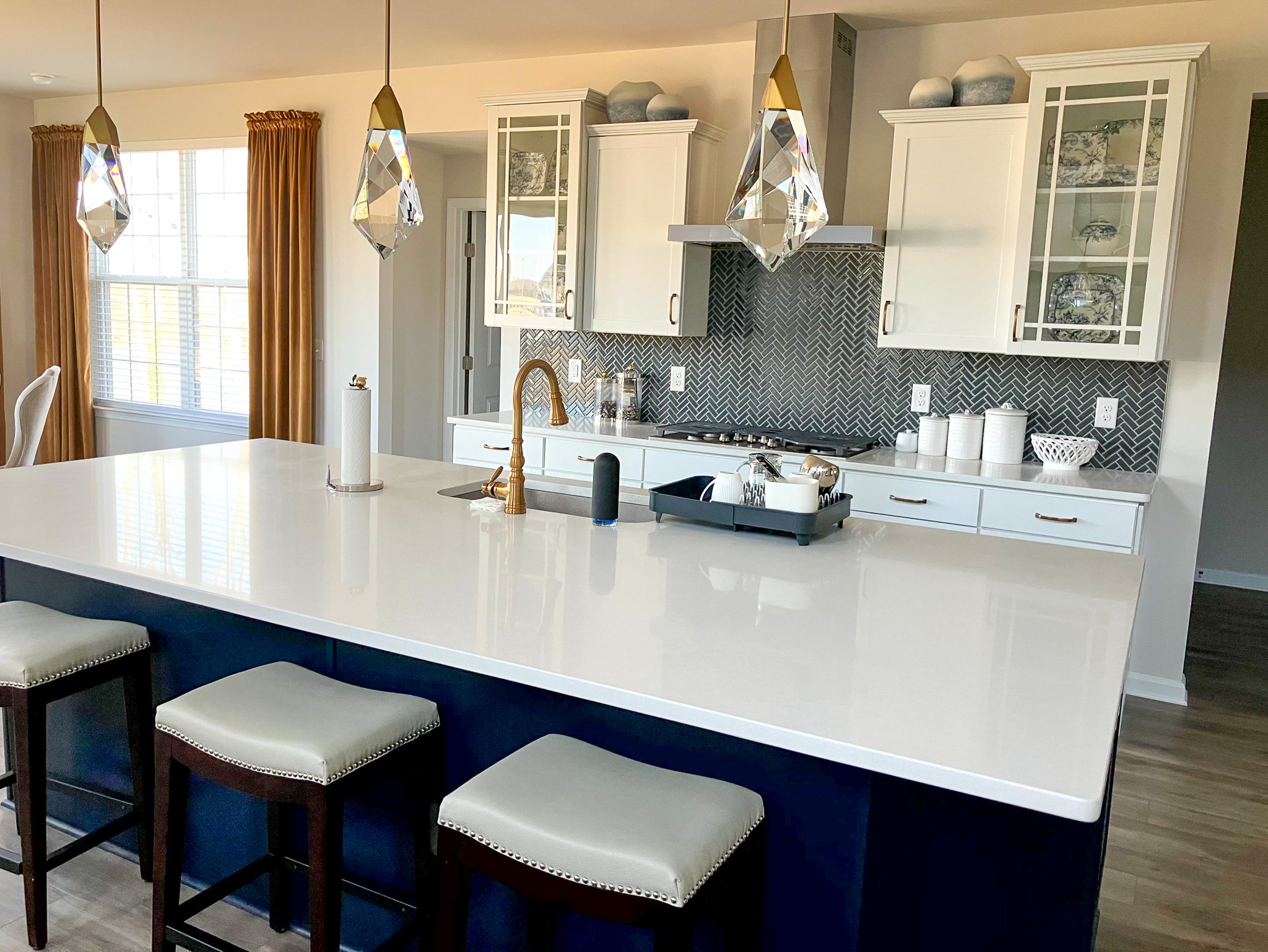 Modern kitchen with a large white island, gold faucet, pendant lights, navy base cabinets, and white upper cabinets with glass doors  inside the home of Sponsored Residential Provider Homaira Yazdani in Fredericksburg, Virginia.