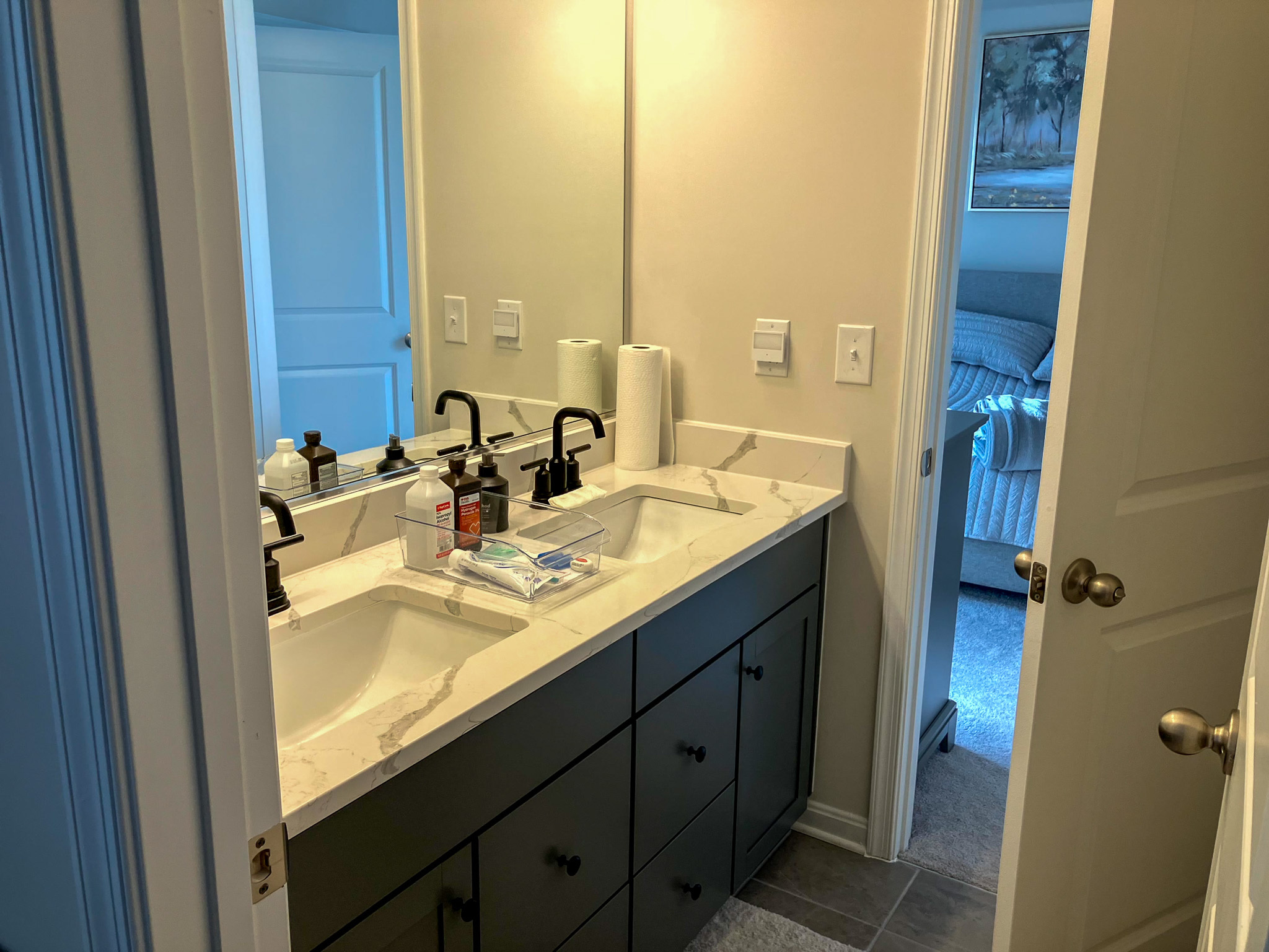 Bathroom with a double-sink vanity, black fixtures, a large mirror, and an open door leading to a bedroom  inside the home of Sponsored Residential Provider Homaira Yazdani in Fredericksburg, Virginia.