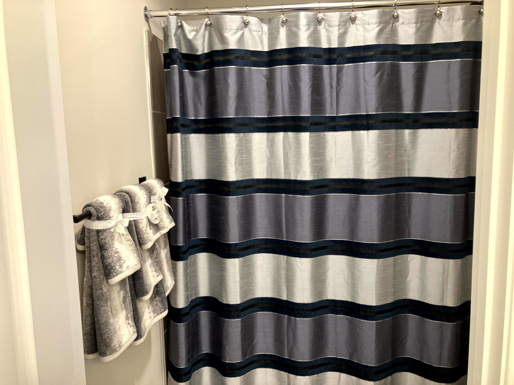 Bathroom with a striped shower curtain in gray, blue, and navy, and folded gray towels hanging on the wall  inside the home of Sponsored Residential Provider Homaira Yazdani in Fredericksburg, Virginia.