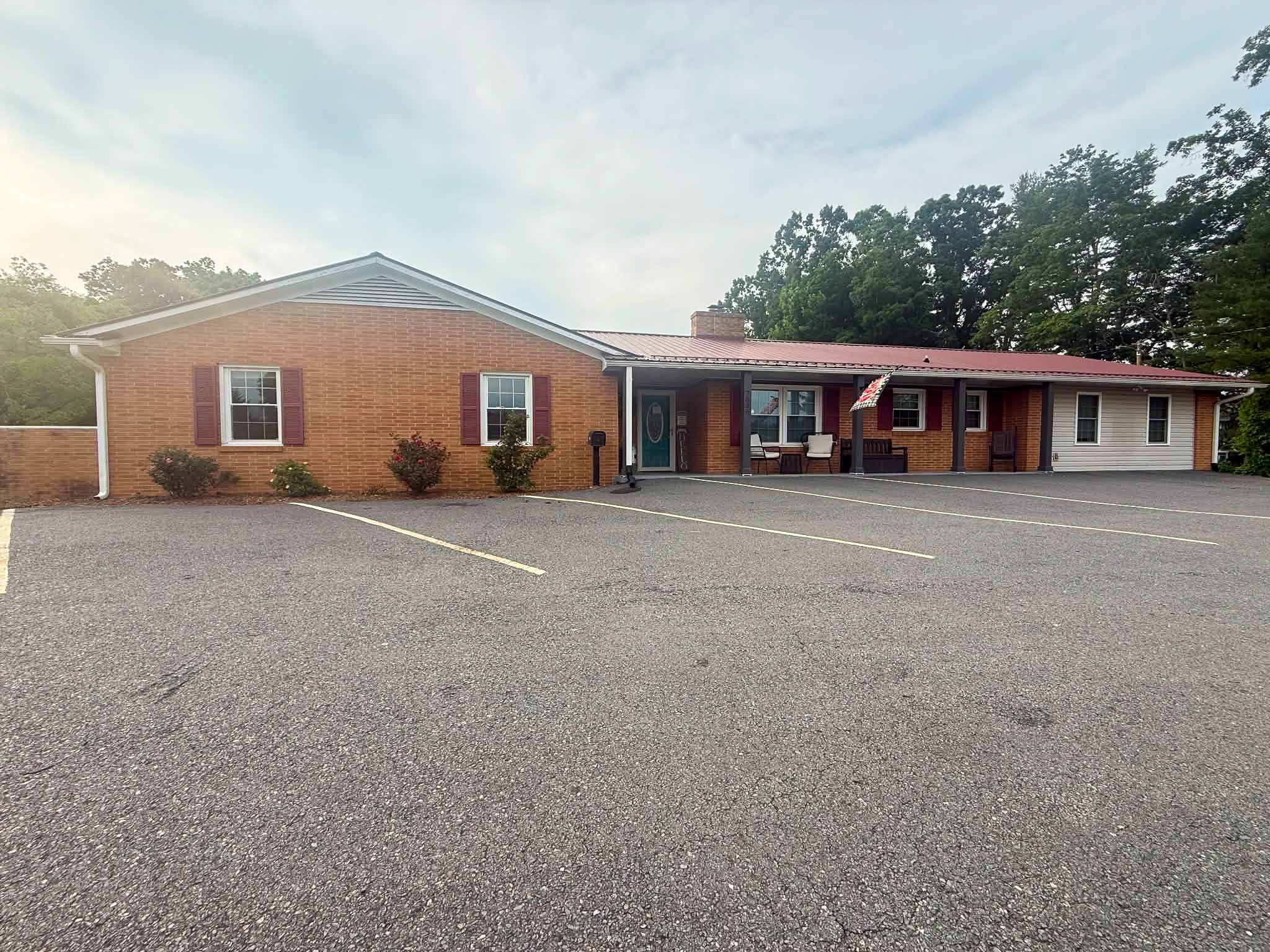 Front of single-story brick building with covered porch seating, red metal roof, and empty parking lot belonging to Group Home Provider Dylan in Galax, Virginia