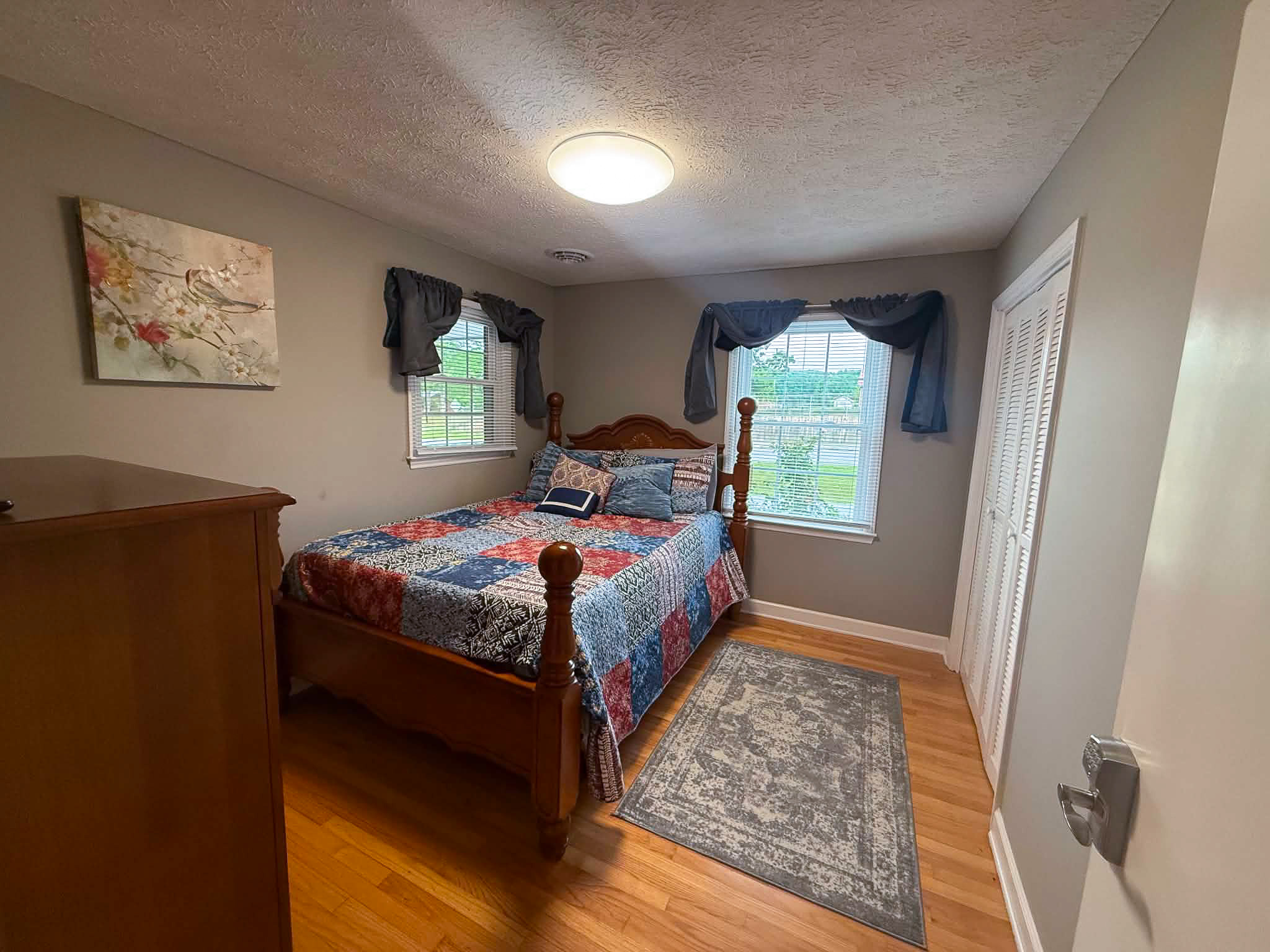 Cozy bedroom with wooden bed frame, patterned quilt, two windows with dark curtains, and hardwood floors inside the home of Group Home Provider Dylan in Galax, Virginia