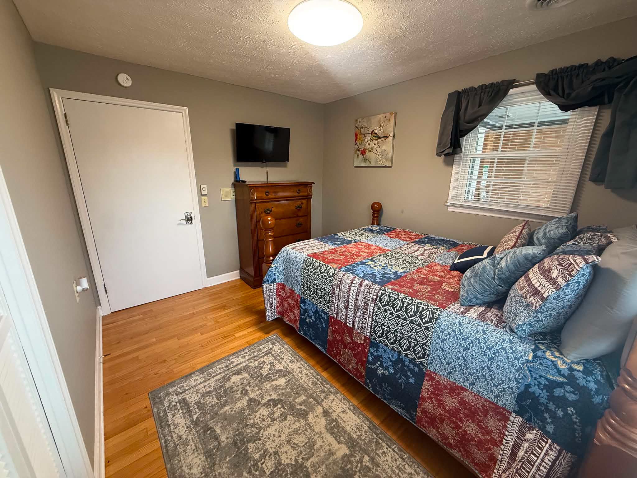 Small bedroom with wooden bed, patterned quilt, wall-mounted TV above dresser, and window with curtains inside the home of Group Home Providers Dylan in Galax, Virginia