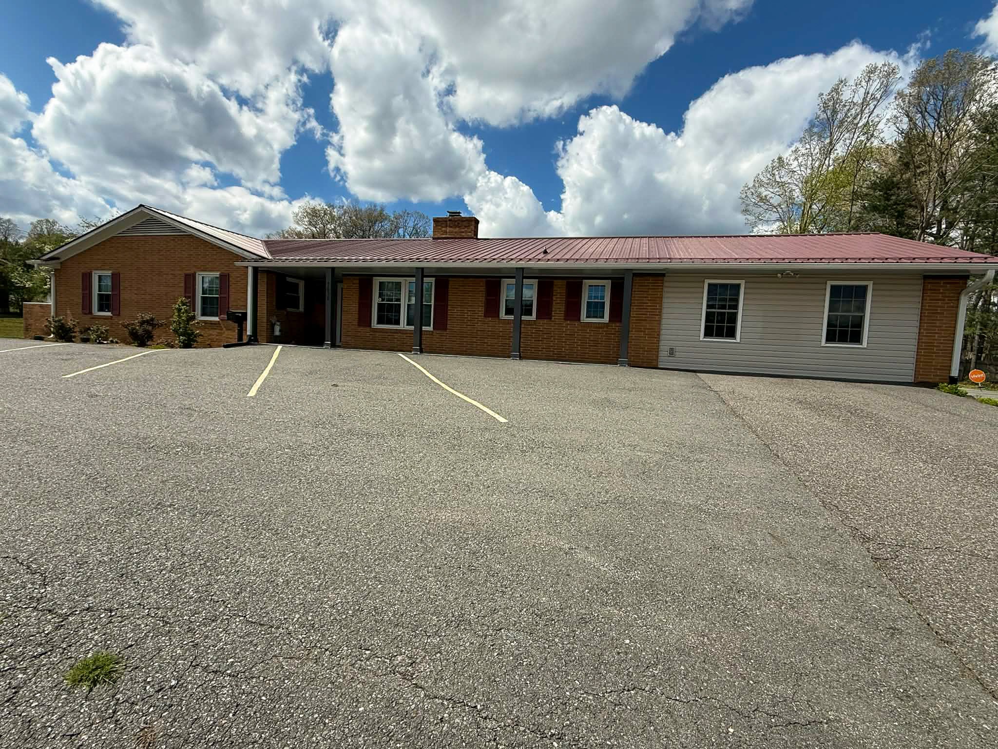 Front view of single-story brick building with covered porch, red shutters, and empty parking lot under blue sky outside the home of Group Home Providers Dylan in Galax, Virginia