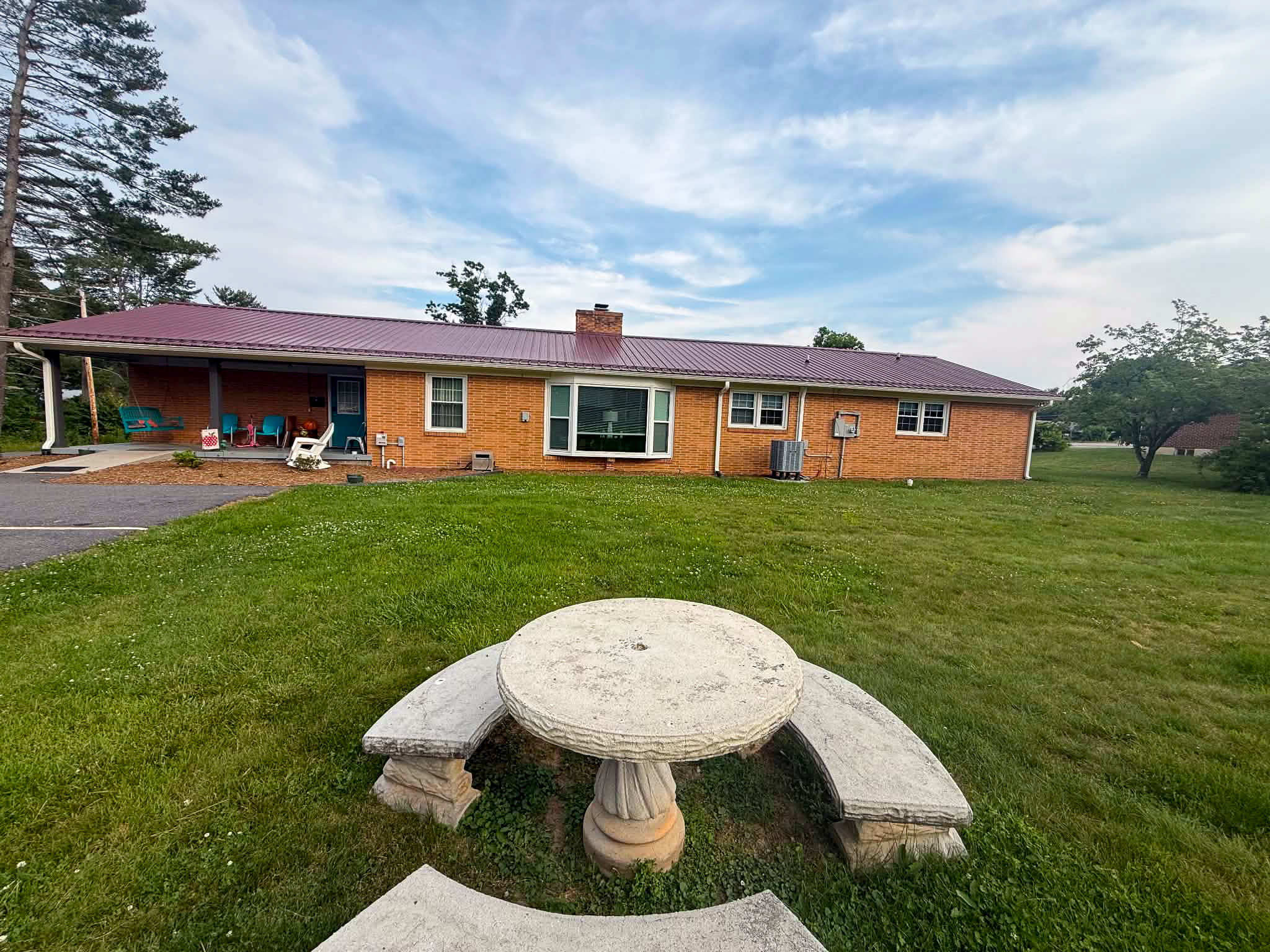 Backyard view of brick home with covered patio, red metal roof, and stone picnic table in grassy yard belonging to Group Home Provider Dylan in Galax, Virginia