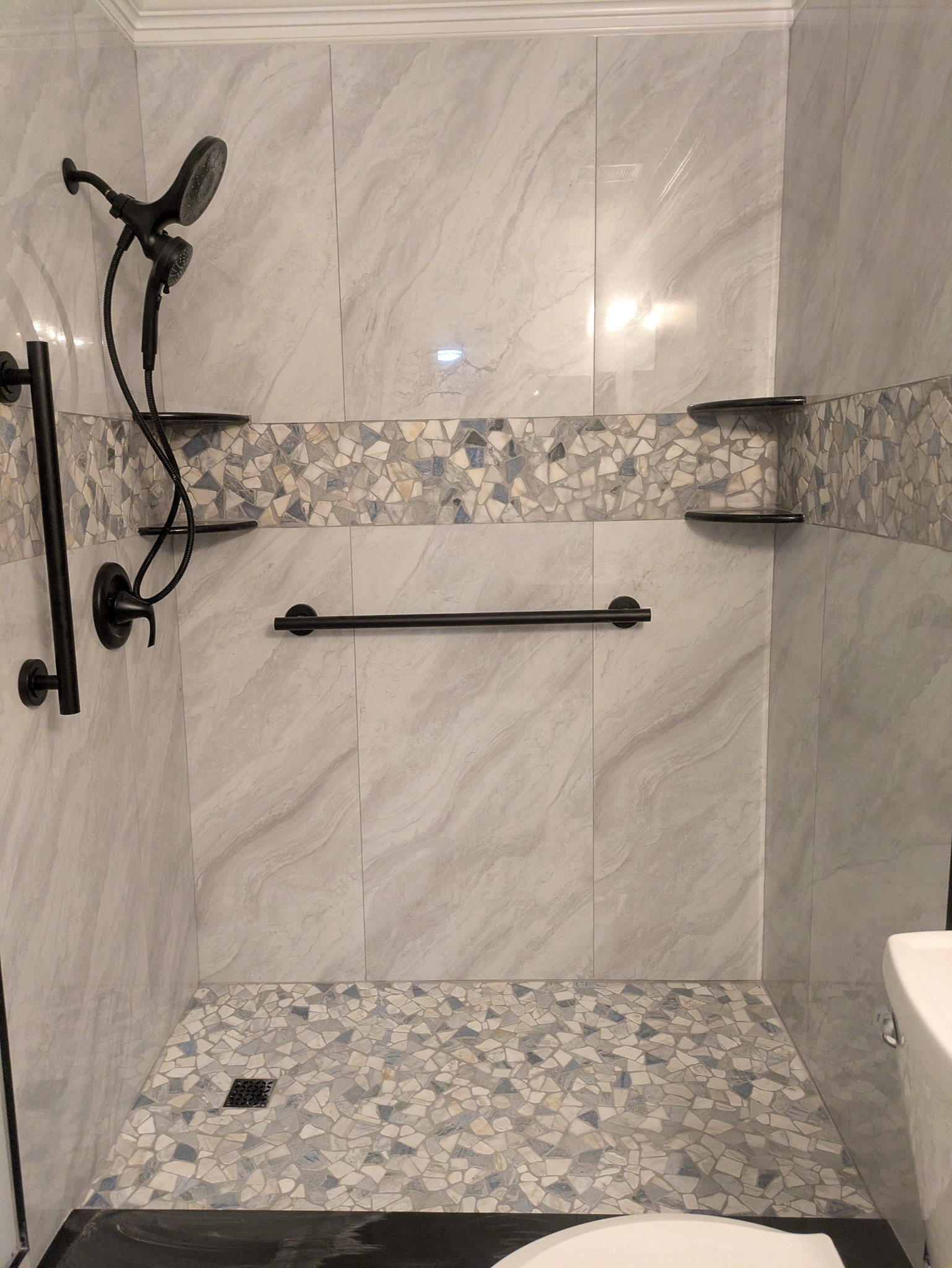 Modern tiled shower with marble-look walls, mosaic accent band, matte black fixtures, built-in corner shelves, and a handheld shower head.