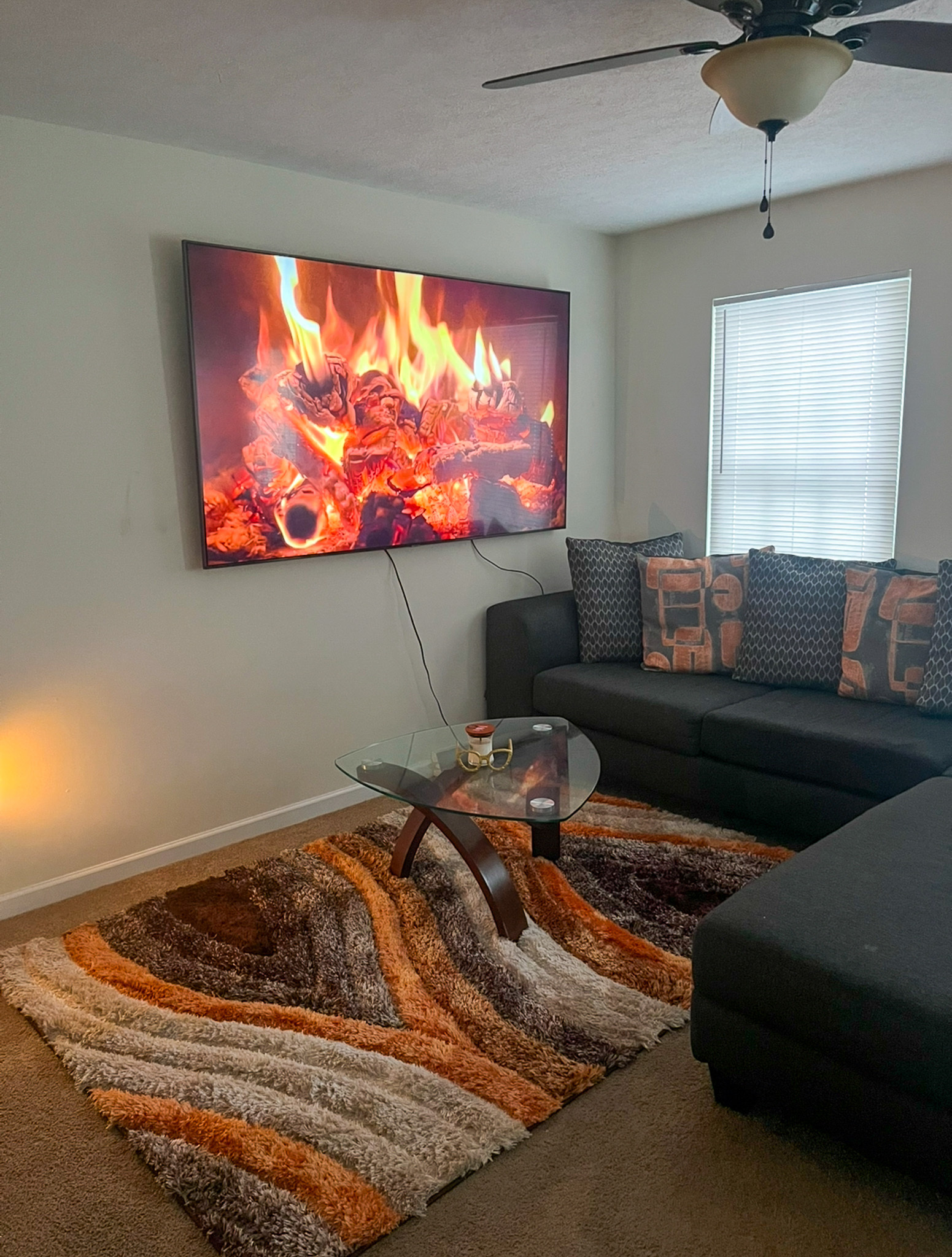 Living room with a sectional sofa, patterned rug, and a wall‑mounted TV displaying a burning fireplace inside the home of Sponsored Residential Provider Kanesha Moseley in Lynchburg, Virginia