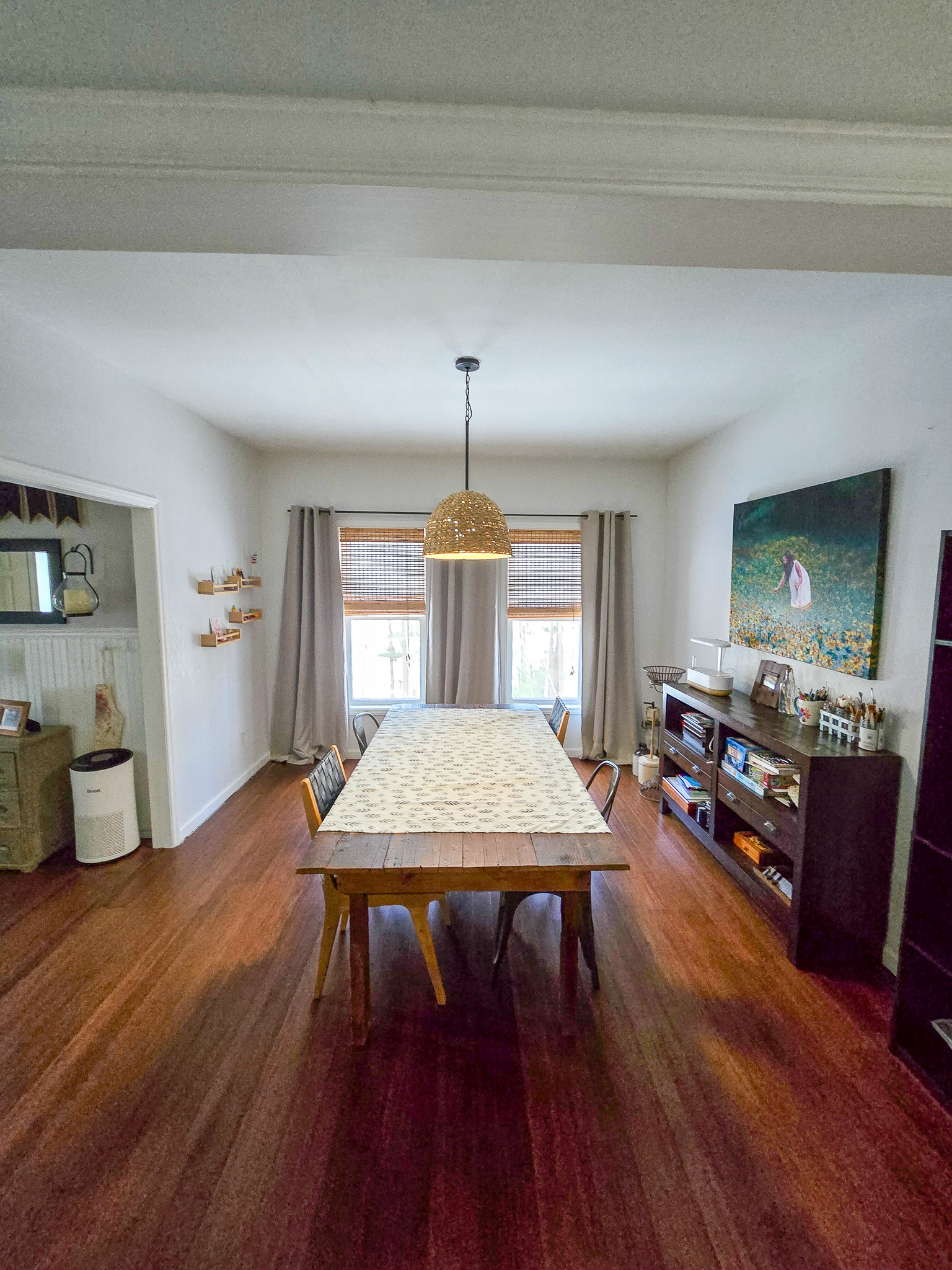A bright dining room with a long table covered by a patterned tablecloth, wooden chairs, a woven pendant light overhead, and tall windows with curtains. Shelving and décor items line the side walls on hardwood floors inside the home of Sponsored Residential Provider Chris Sumner in Galax, Virginia.