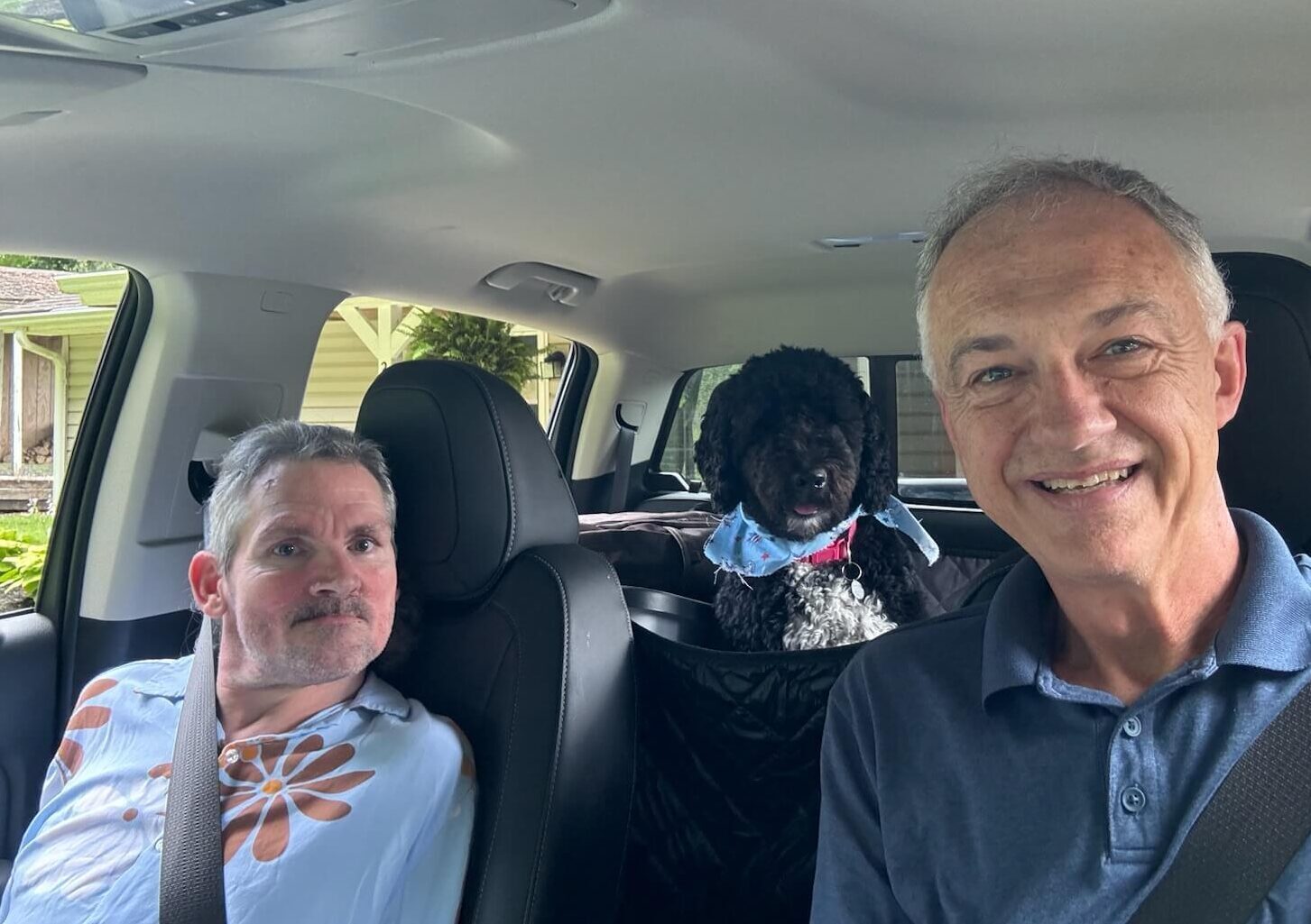 Two men taking a selfie in the car with a black dog in the back seat