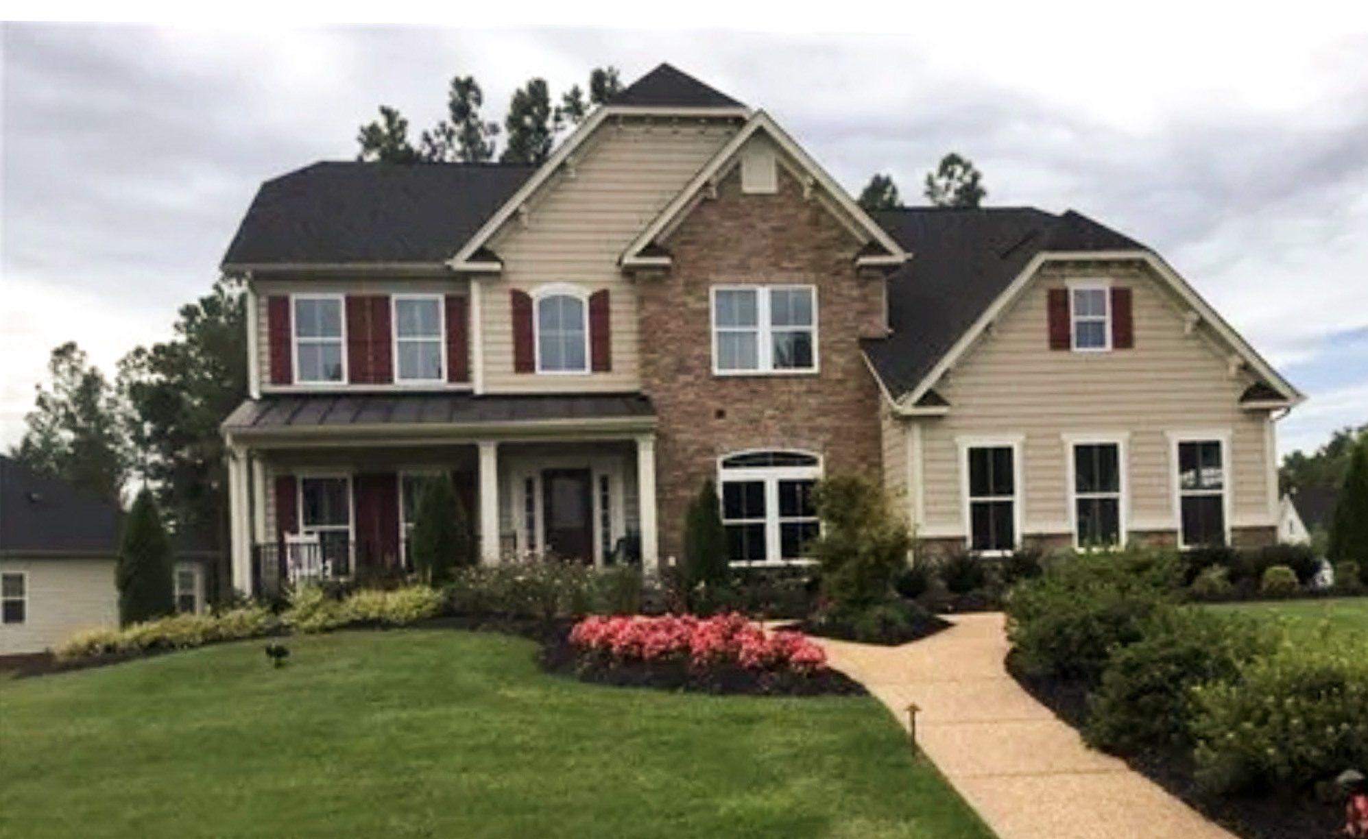 Two-story suburban house with beige siding and stone facade, black roof, red shutters, landscaped lawn, and curved front walkway belonging to group home providers Sheniqua and Travon Jackson in Richmond, VA.