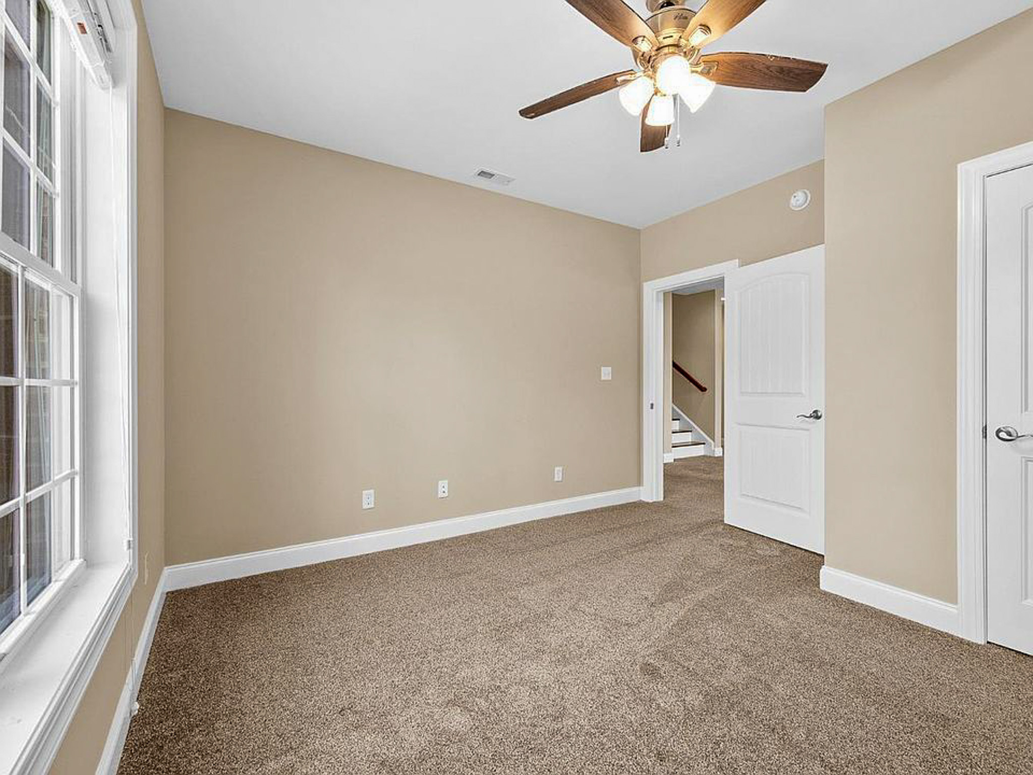 Empty, carpeted available bedroom with beige walls, ceiling fan, and large window, in the home of sponsored residential provider Lisa Reed in Lynchburg, Virginia.
