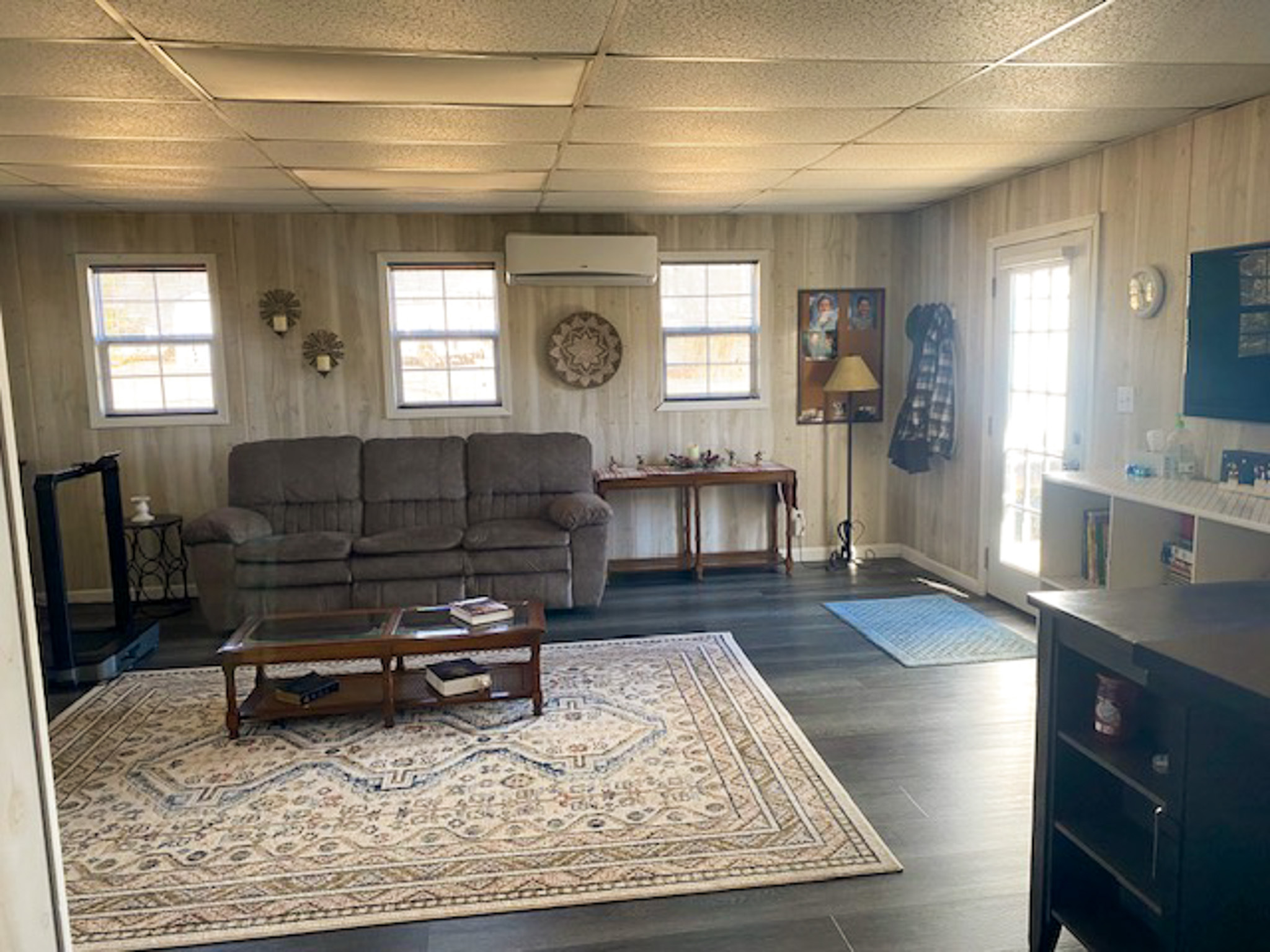 A cozy living area with wood‑panel–style walls, a gray reclining sofa, a patterned rug on dark flooring, and several windows letting in light beside a door to the right inside the home of Sponsored Residential Provider Christina Bedsaul in Hillsville, Virginia.