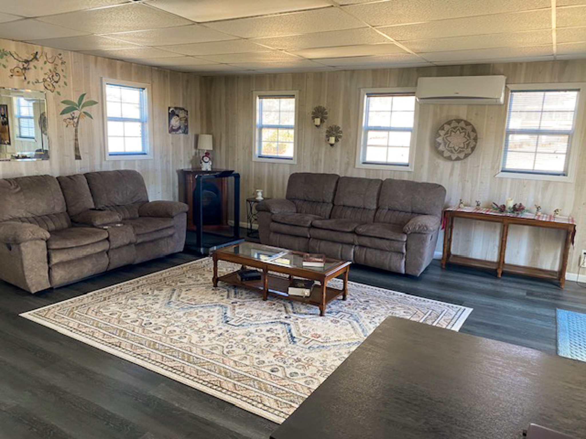A spacious living room with two brown sofas, a patterned area rug on dark flooring, wood‑panel–style walls, and several windows providing natural light, with a console table and wall décor along the back wall inside the home of Sponsored Residential Provider Christina Bedsaul in Hillsville, Virginia.