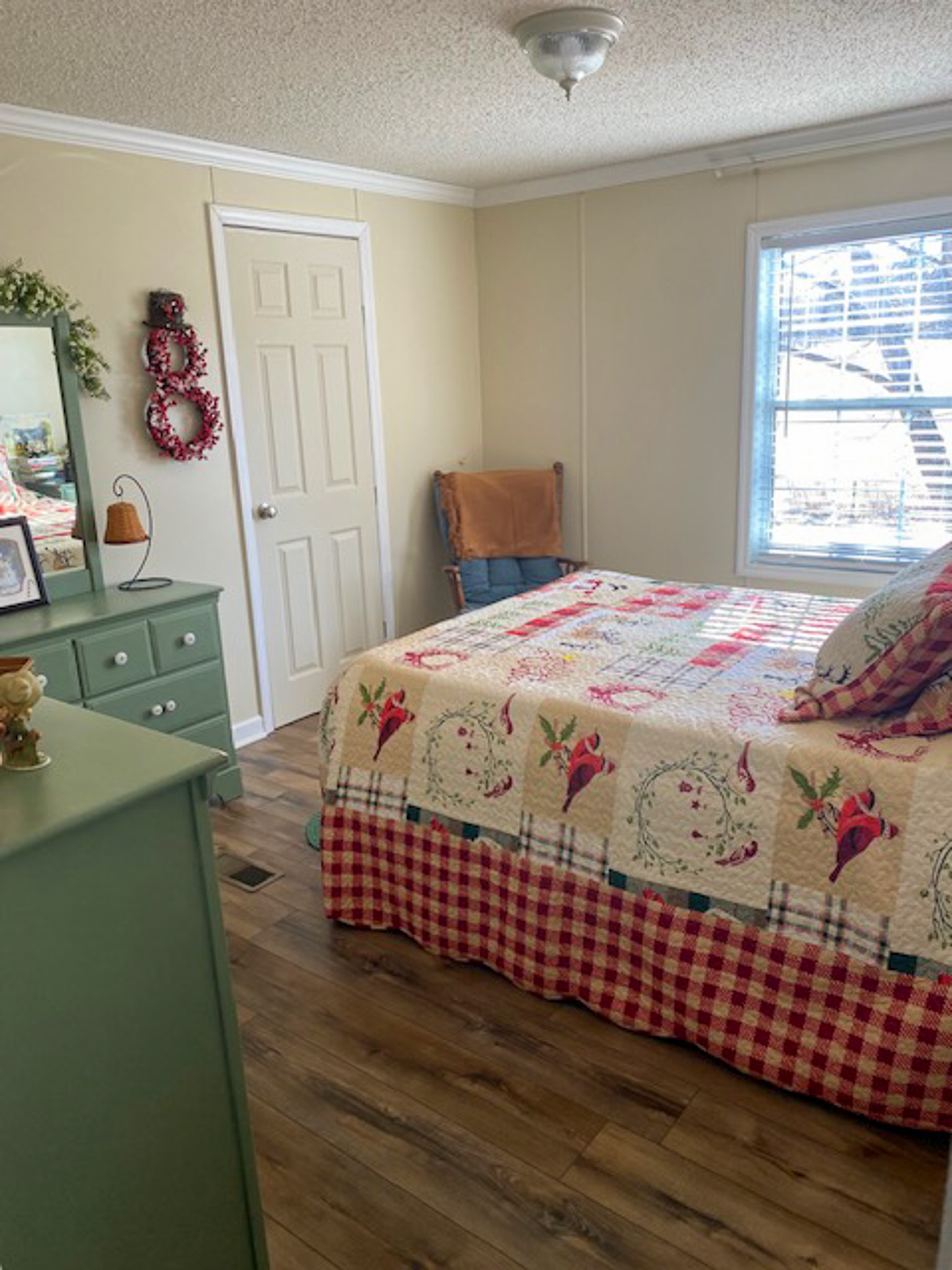 A bright bedroom with a patterned quilt on the bed, wood‑look flooring, a green dresser with a mirror, wall décor, and a window letting in natural light near a closed white door inside the home of Sponsored Residential Provider Christina Bedsaul in Hillsville, Virginia.
