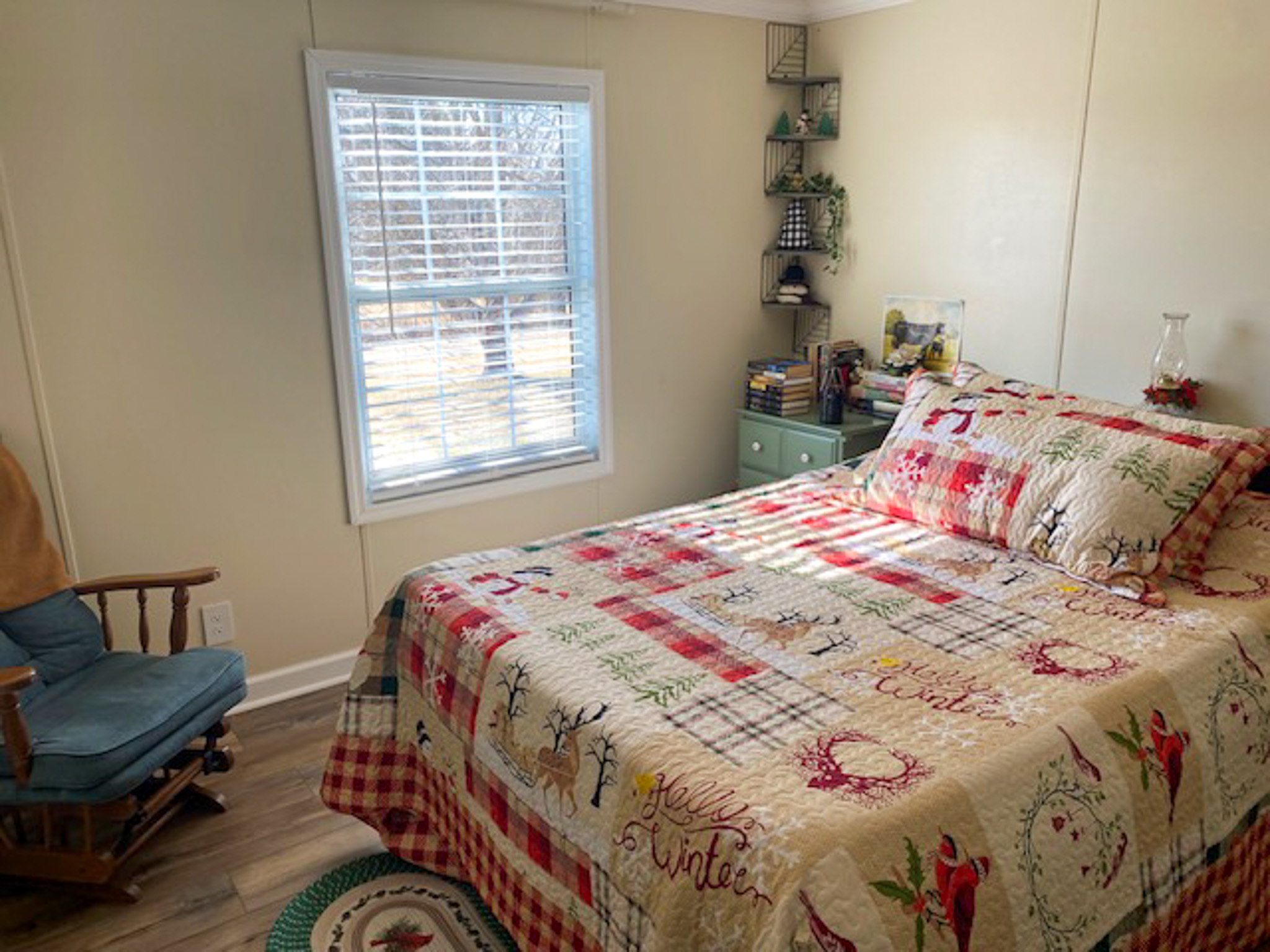 A small, bright bedroom with a bed dressed in a colorful patterned quilt, a window letting in natural light, a wooden rocking chair, and a corner shelf above a green dresser inside the home of Sponsored Residential Provider Christina Bedsaul in Hillsville, Virginia.