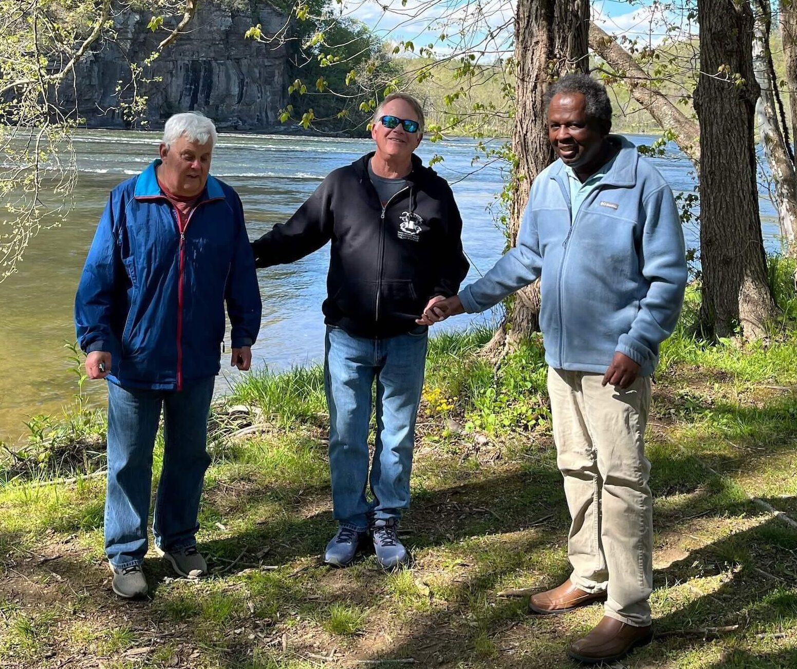 Three men standing together in front of a lake and trees