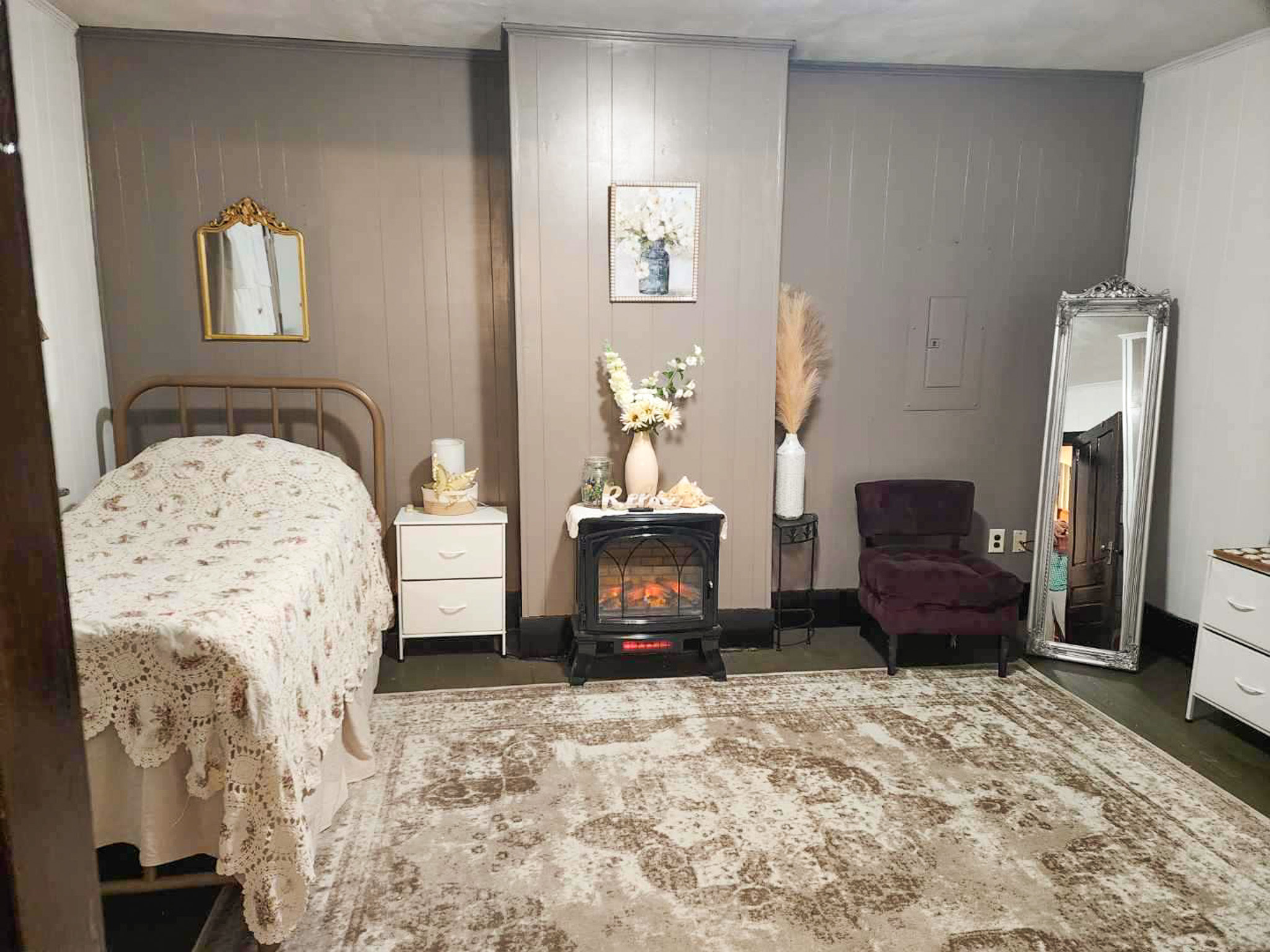 Bedroom with a floral bedspread, two white nightstands, an electric fireplace, a purple chair, and a tall mirror on a patterned rug inside the home of Sponsored Residential Provider Angela Justice in Mathews, Virginia.
