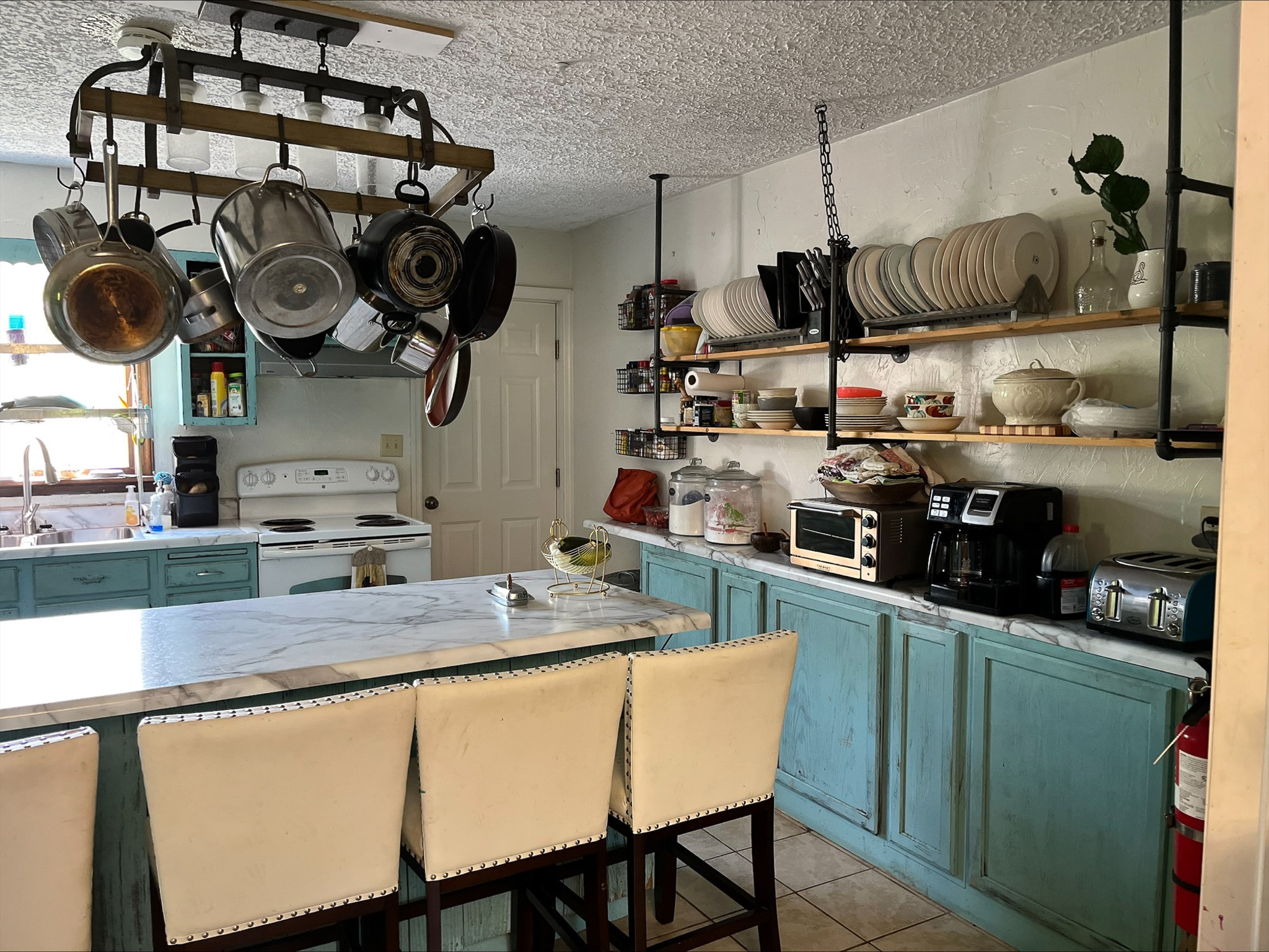 Kitchen with turquoise cabinets, marble‑top island, hanging pot rack, and open shelving filled with dishes and small appliances inside the home of Sponsored Residential Provider Dara Short in Willis, Virginia.