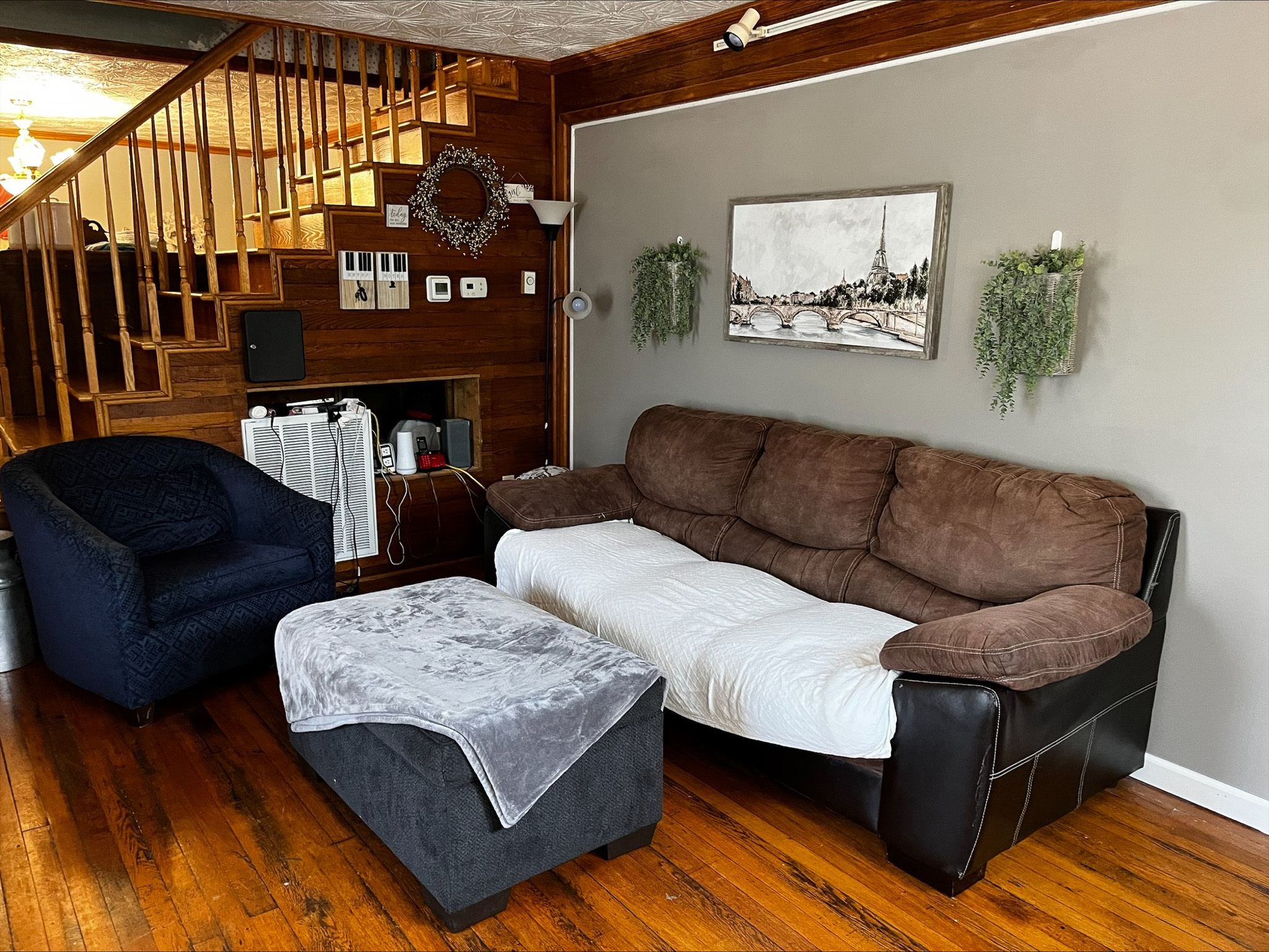 Living room with a brown and black sofa, blue armchair, large ottoman, wood floors, and a staircase with wooden railings in the background inside the home of Sponsored Residential Provider Dara Short in Willis, Virginia.