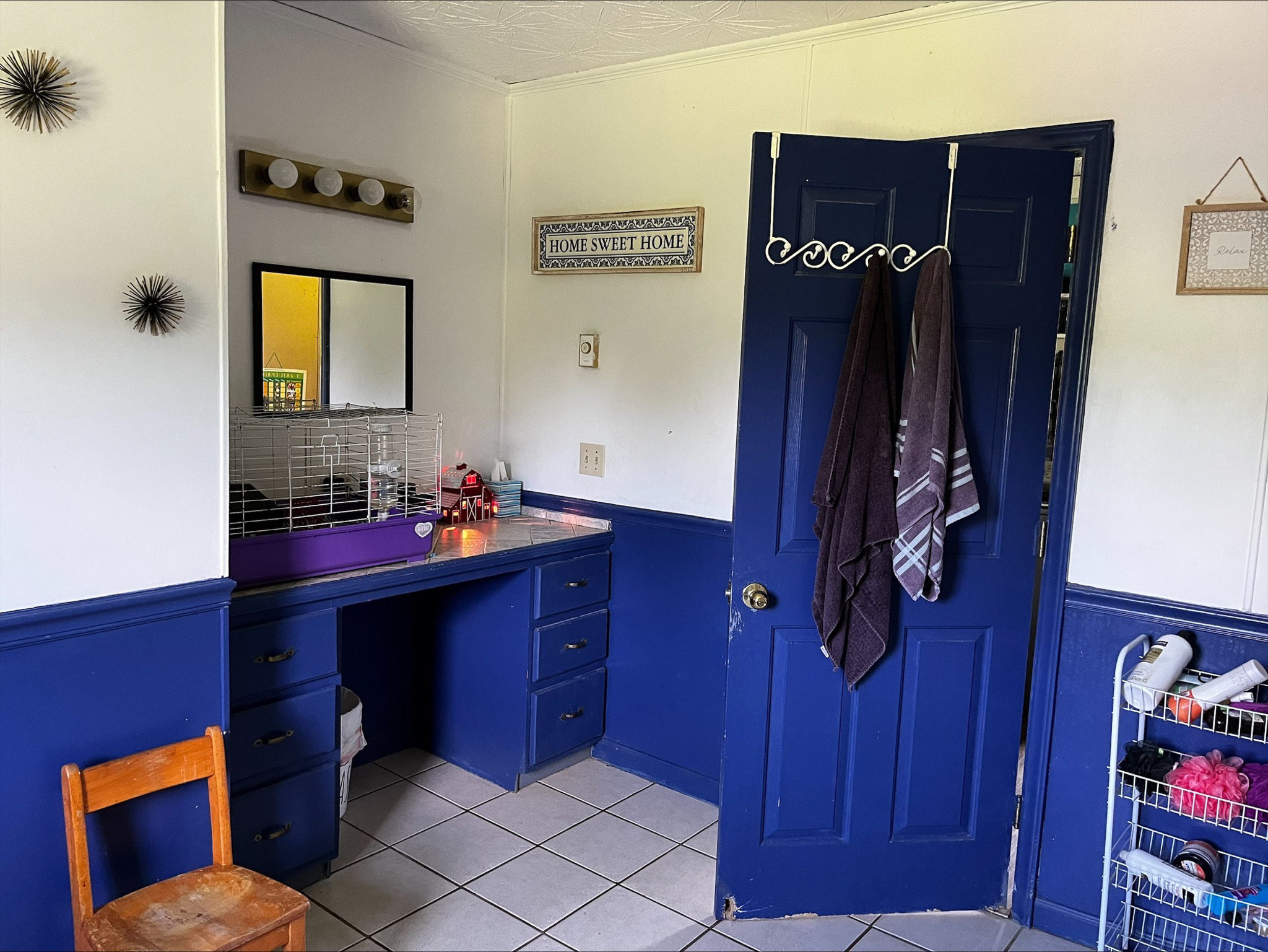 Bathroom with blue lower walls and cabinetry, a vanity with mirror and lights, a blue door with towels hanging on hooks, and tiled flooring inside the home of Sponsored Residential Provider Dara Short in Willis, Virginia.