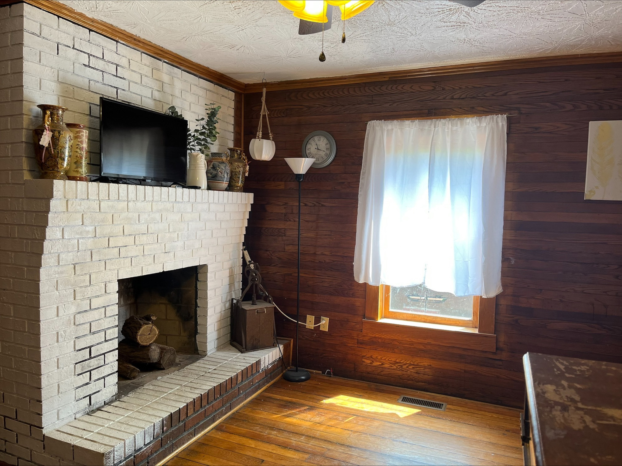 Room with a white-painted brick fireplace, wood-paneled walls, a window with white curtains, and hardwood floors inside the home of Sponsored Residential Provider Dara Short in Willis, Virginia.