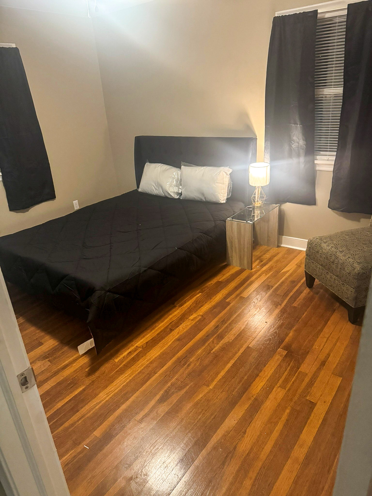 A small bedroom with a dark‑colored bed, a bedside lamp on a light wood nightstand, dark curtains, hardwood floors, and a patterned armchair in the corner inside the home of Sponsored Residential Provider Selena Banks in Chesterfield, Virginia.