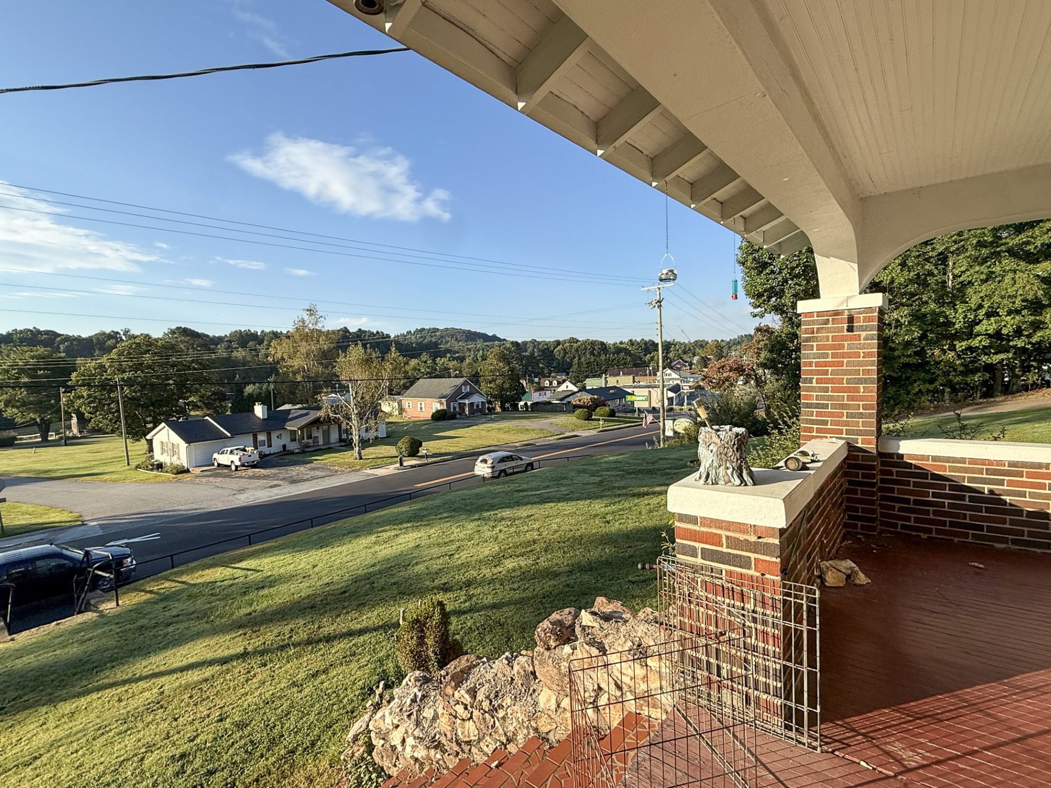 Sunny view from a brick front porch overlooking a grassy yard, road, and houses with hills in the distance at the home of Sponsored Residential Provider Tammy Hunt in Floyd, Virginia.