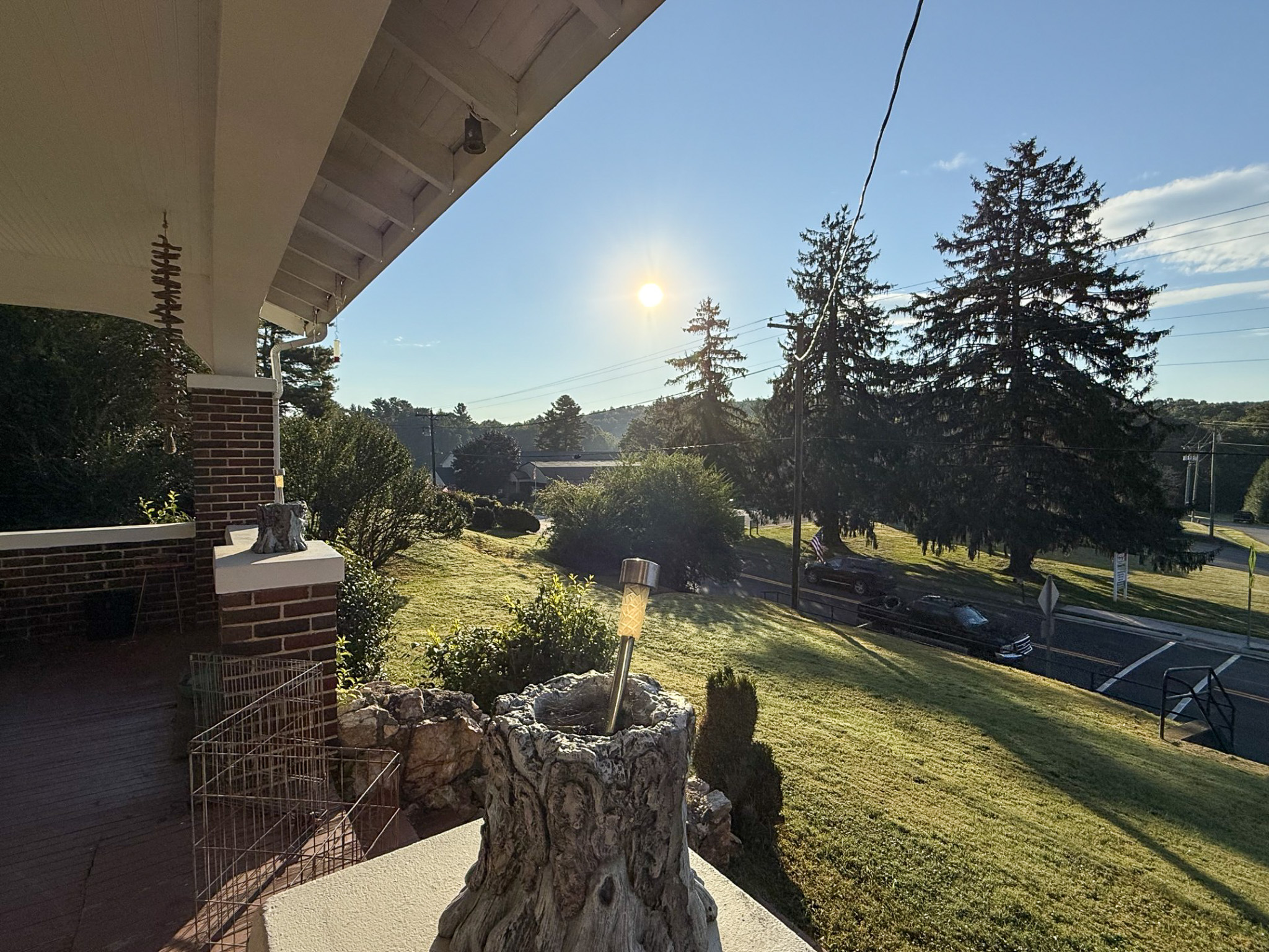 Sunlit view from a brick porch overlooking a grassy slope, trees, and a road, with the sun low in the sky from the front porch of the home of Sponsored Residential Provider Tammy Hunt in Floyd, Virginia.